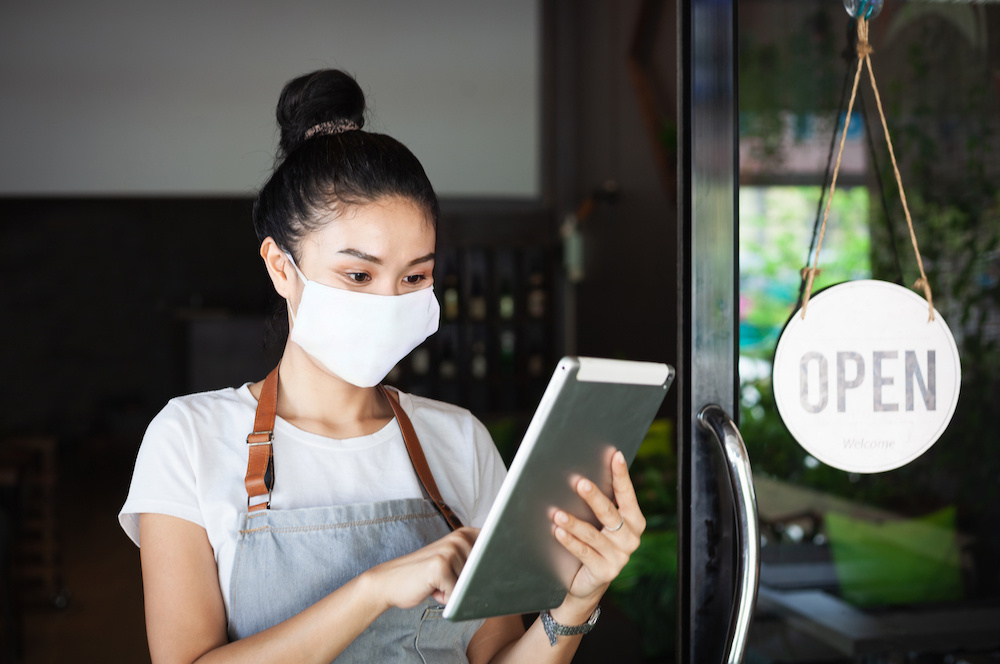 SME entrepreneur in a mask checking off items on a tablet as store opens